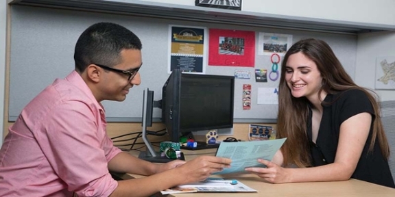 Two people sitting at a desk discussing a pamphlet in front of them