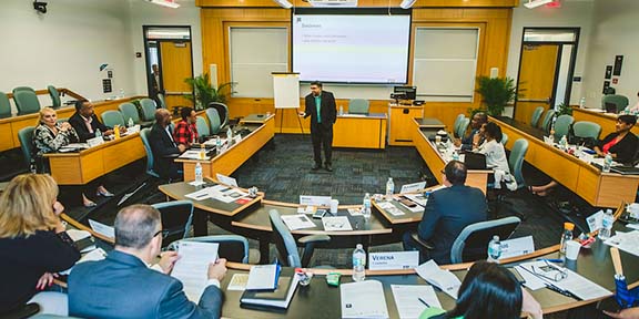 A lecturer standing at the front of the classroom as the students listen and take notes around him. 